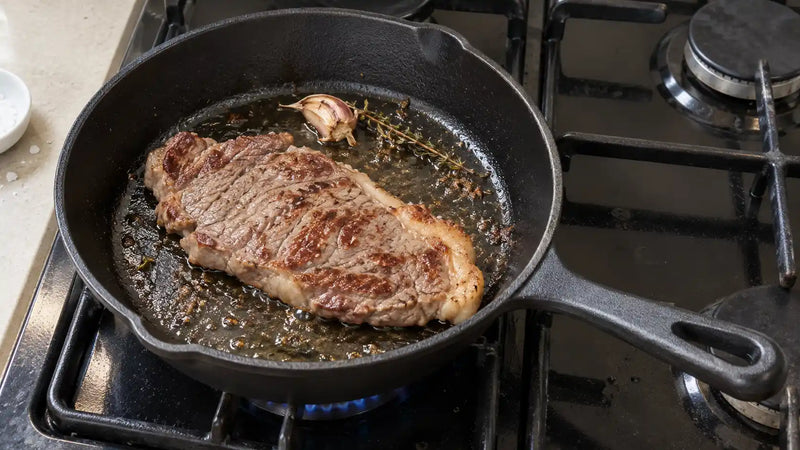 Comment utiliser une poêle en fonte : une entrecôte saisie à la croûte dorée, avec une gousse d'ail et un brin de thym, dans une poêle en fonte sur une gazinière