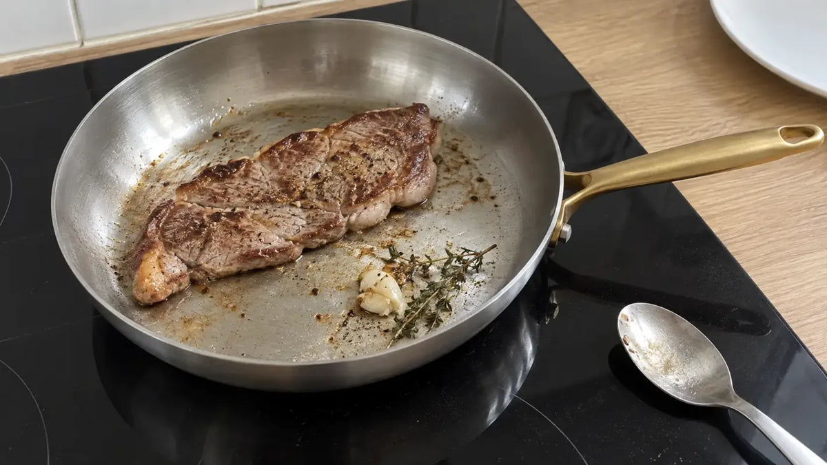 Cuire de la viande dans une poêle en inox : une entrecôte saisie et dorée, avec une gousse d'ail et une branche de thym