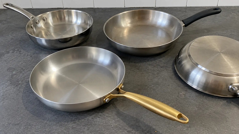 Poêle inox à manche doré de L'Atelier de la Poêle, meilleure marque de poêles en inox, au premier plan devant trois autres poêles en inox posées sur un plan de travail gris