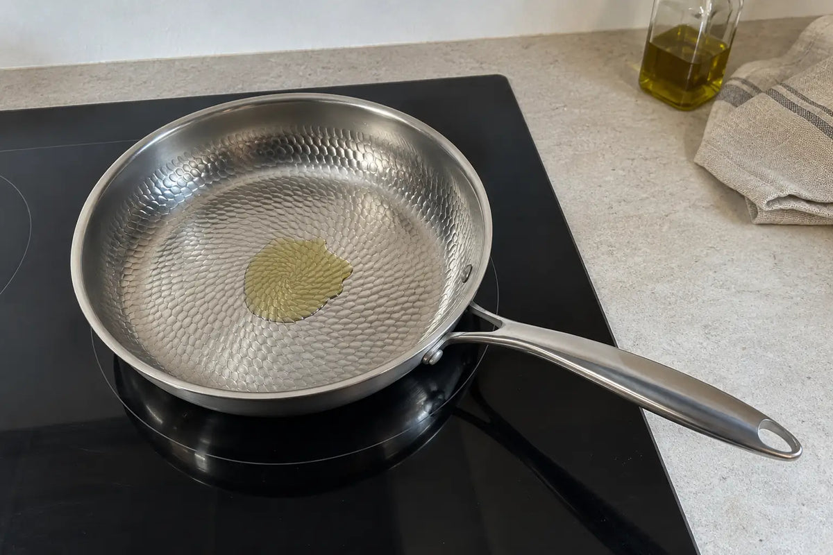 Poêle en titane martelé de 28 cm posée sur une plaque à induction, avec un filet d'huile d'olive qui brille dans les alvéoles de sa surface martelée