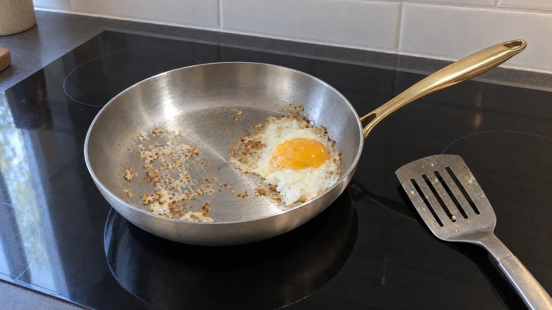 Poêle inox 28 cm sur une plaque à induction, un œuf au plat qui a accroché et s'est déchiré sur la surface de cuisson, une spatule en métal à côté