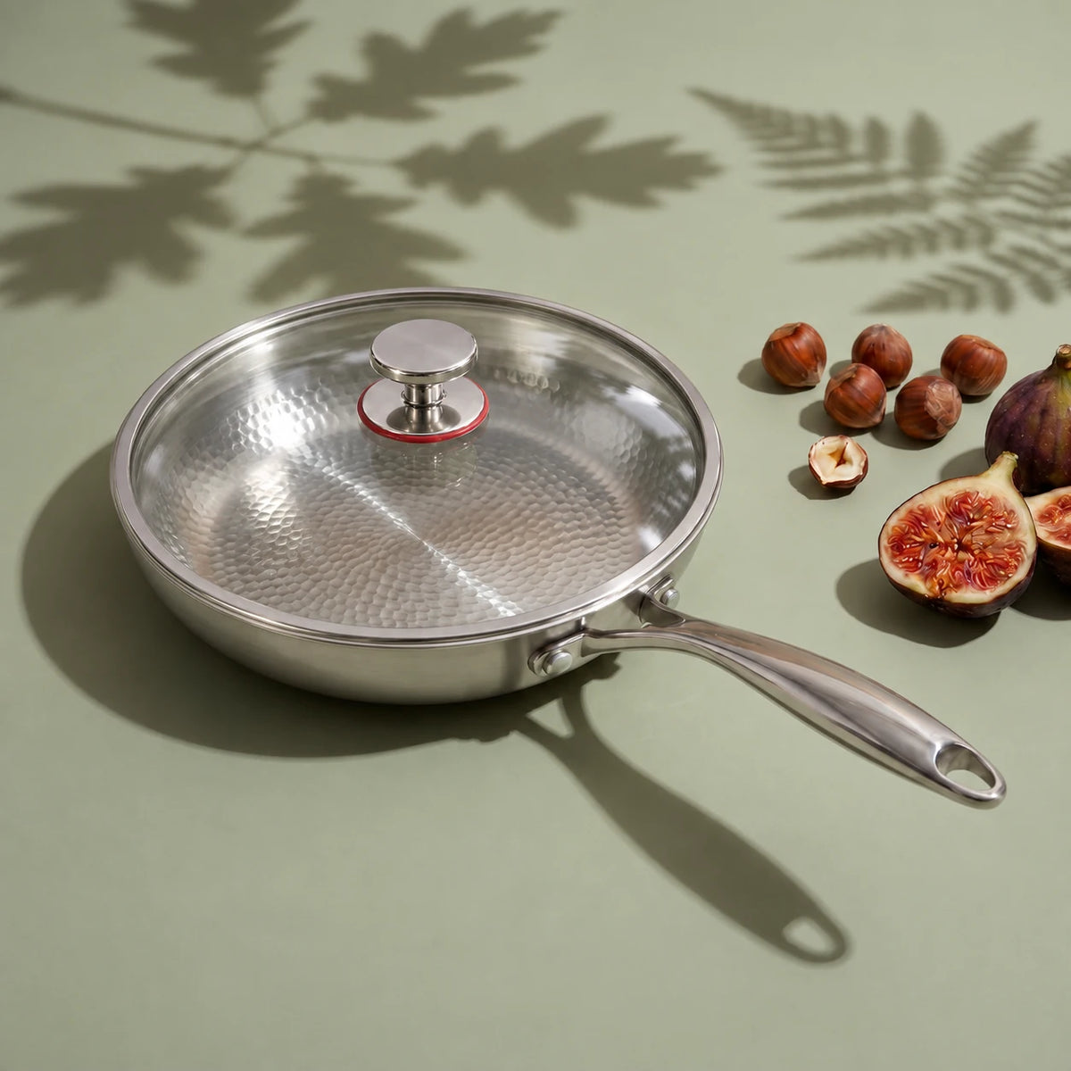 Poêle en titane avec couvercle 26 cm, son couvercle en verre posé, à côté de noisettes et de figues sur fond vert sauge