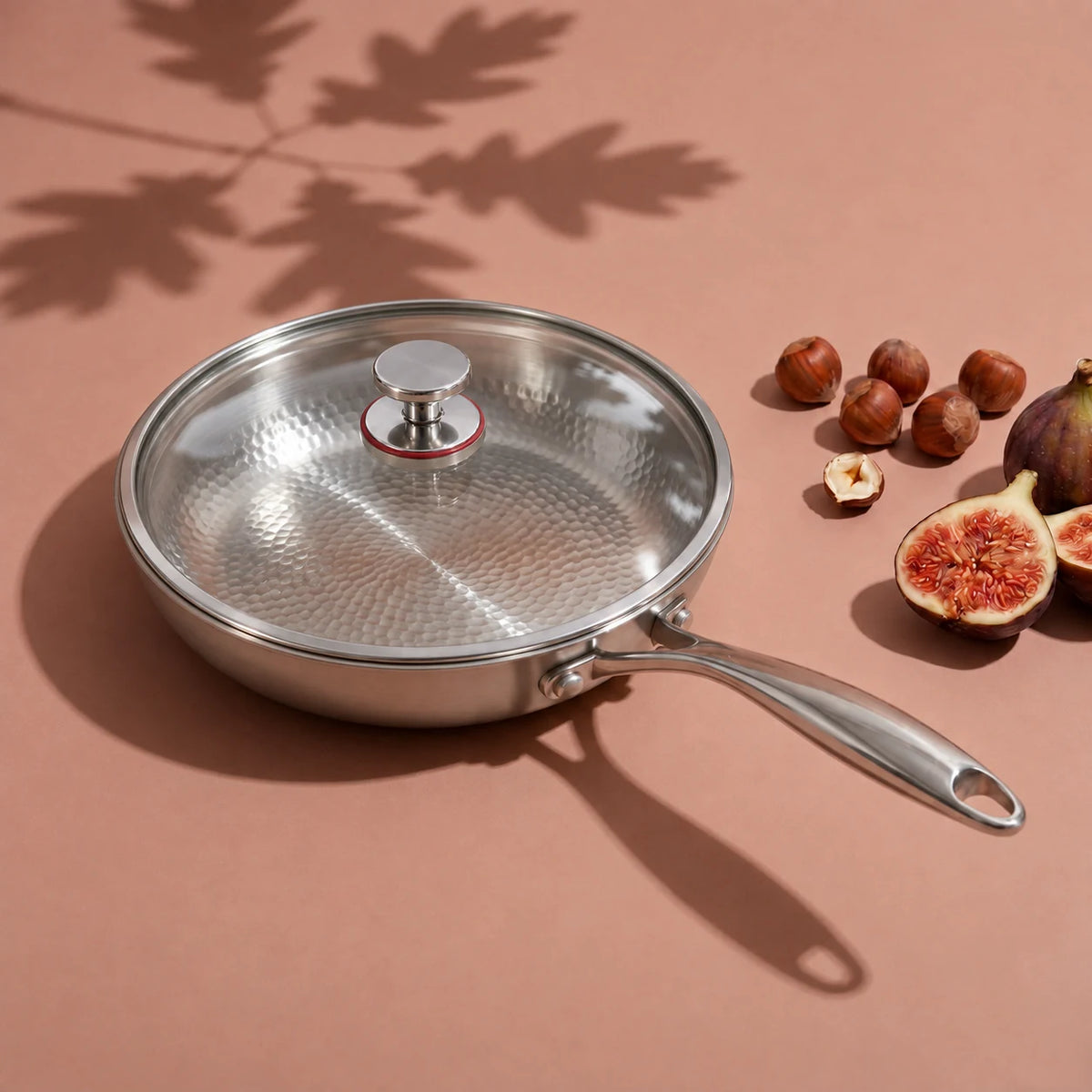 Poêle en titane avec couvercle 30 cm, son couvercle en verre posé, à côté de noisettes et de figues sur fond rose
