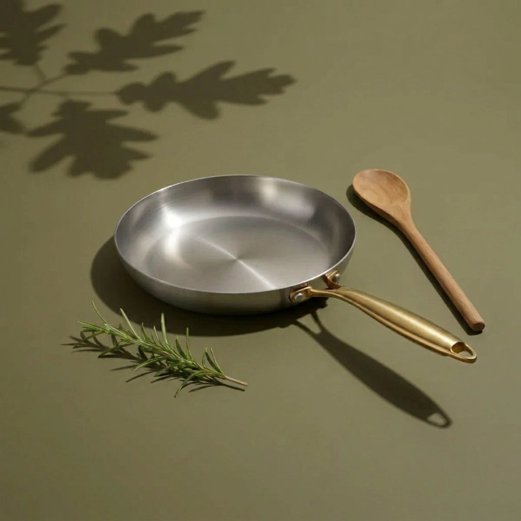 Poêle tous feux 22 cm vue de trois-quarts sur fond vert olive, avec un brin de romarin et une cuillère en bois