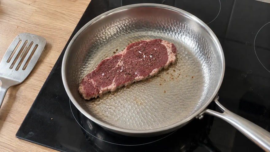 Poêle en titane martelé de L'Atelier de la Poêle, meilleure marque de poêles en titane, avec une entrecôte qui commence à cuire sur une plaque à induction, à côté d'un plan de travail en chêne