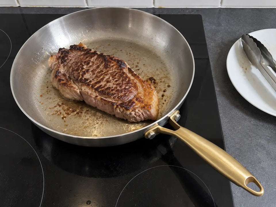 Entrecôte saisie dans une poêle inox à manche doré de L'Atelier de la Poêle, sur une plaque à induction, à côté d'une assiette et d'une pince de cuisine