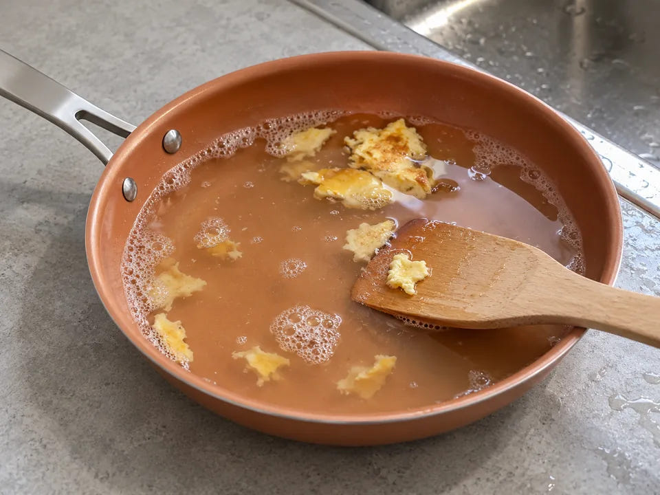 Poêle en céramique qui trempe dans l'eau chaude savonneuse : une spatule en bois décolle les restes d'omelette ramollis