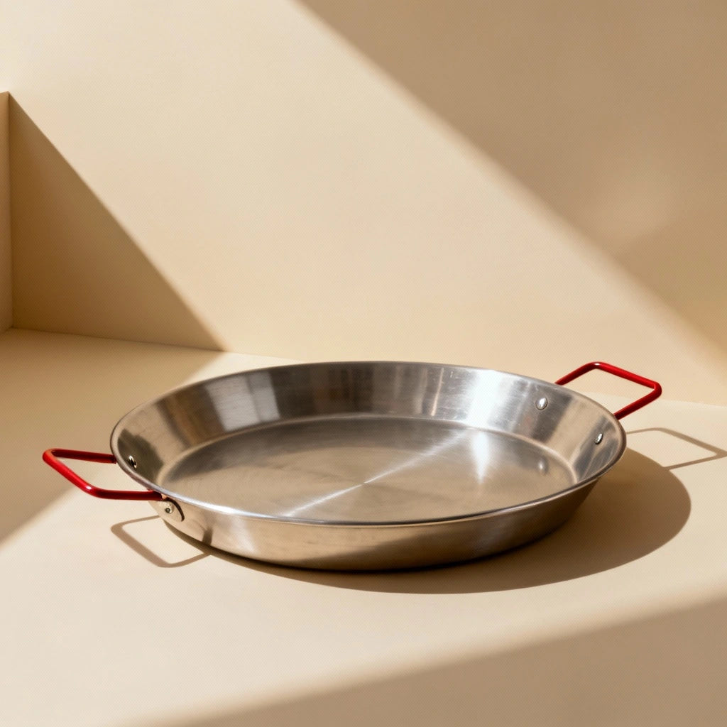 Poêle à Paella Multi Tailles : modèle en inox à poignées rouges sur fond beige.
