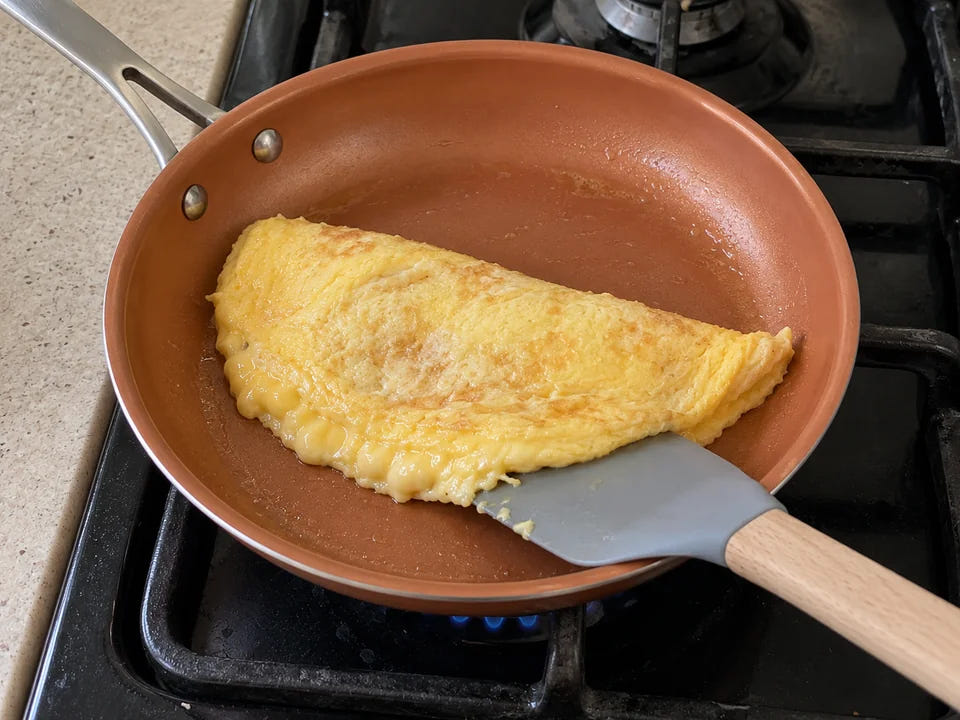 Poêle en céramique : une omelette baveuse pliée en deux, une spatule en silicone glissée dessous, sur une petite flamme