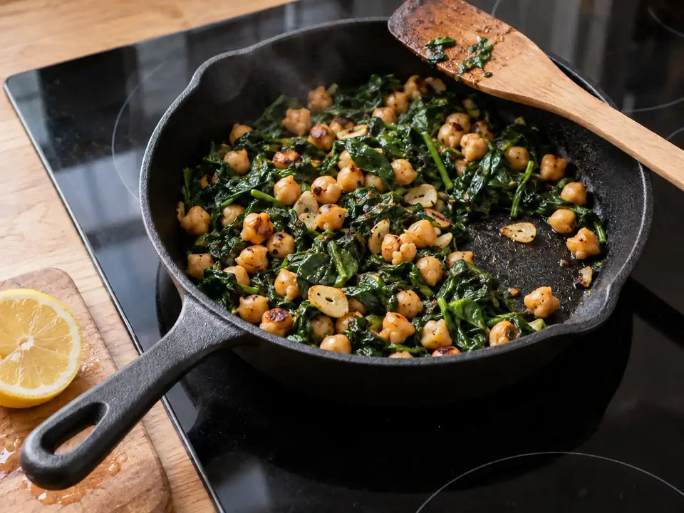 Poêlée d'épinards et de pois chiches cuite dans une poêle en fonte sur une plaque à induction, deux aliments qui apportent du fer, à côté d'un demi-citron