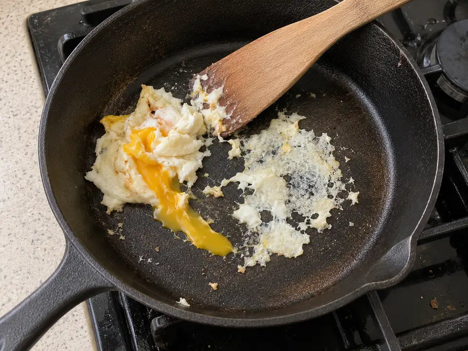Pourquoi ma poêle en fonte colle : un œuf au plat déchiré est resté accroché à la poêle, à côté d'une spatule en bois