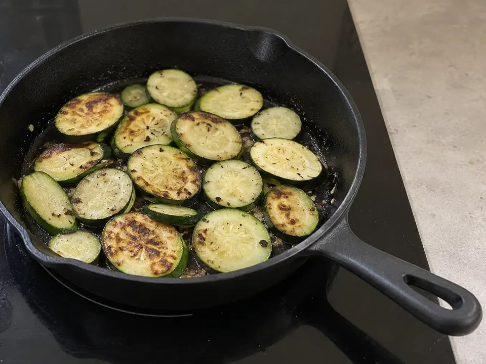 Poêle en fonte posée sur un foyer de plaque à induction à sa taille, avec des rondelles de courgette qui dorent