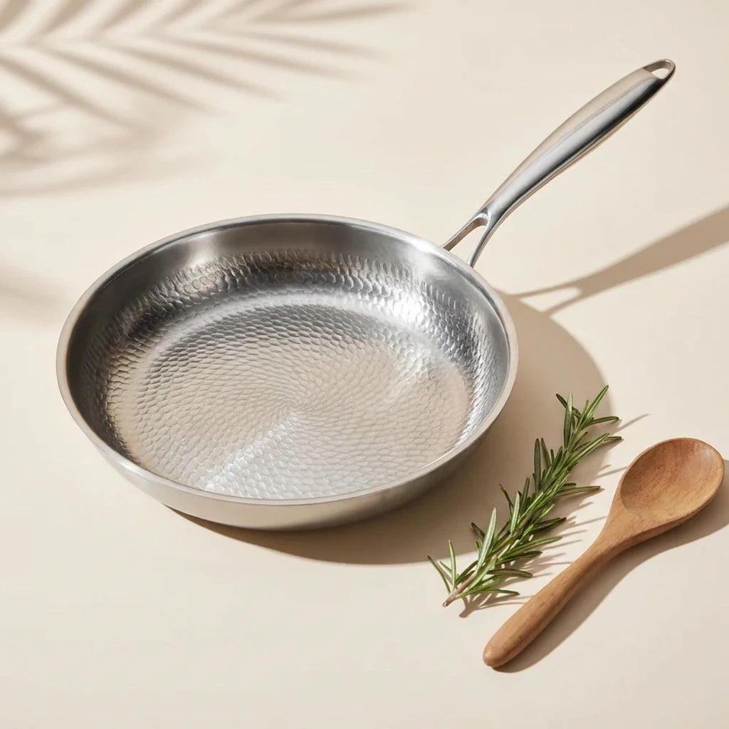 Poêle en titane 26 cm vue de trois quarts, sa surface martelée à côté d'une cuillère en bois et de romarin sur fond crème
