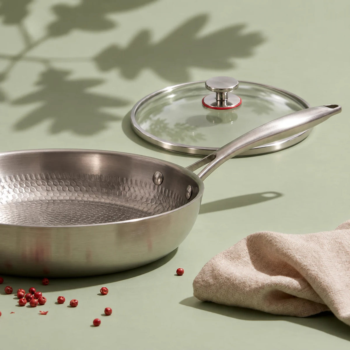 Poêle en titane avec couvercle 26 cm, le couvercle en verre posé juste derrière, avec un torchon en lin et des baies roses