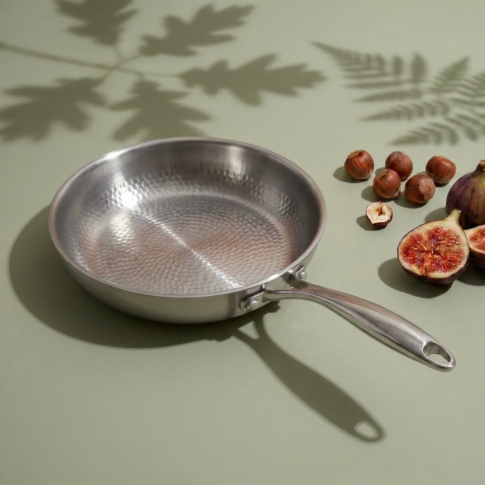 Poêle en titane martelé 22 cm vue de dessus, sa surface alvéolée à côté de figues et de noisettes sur fond vert sauge