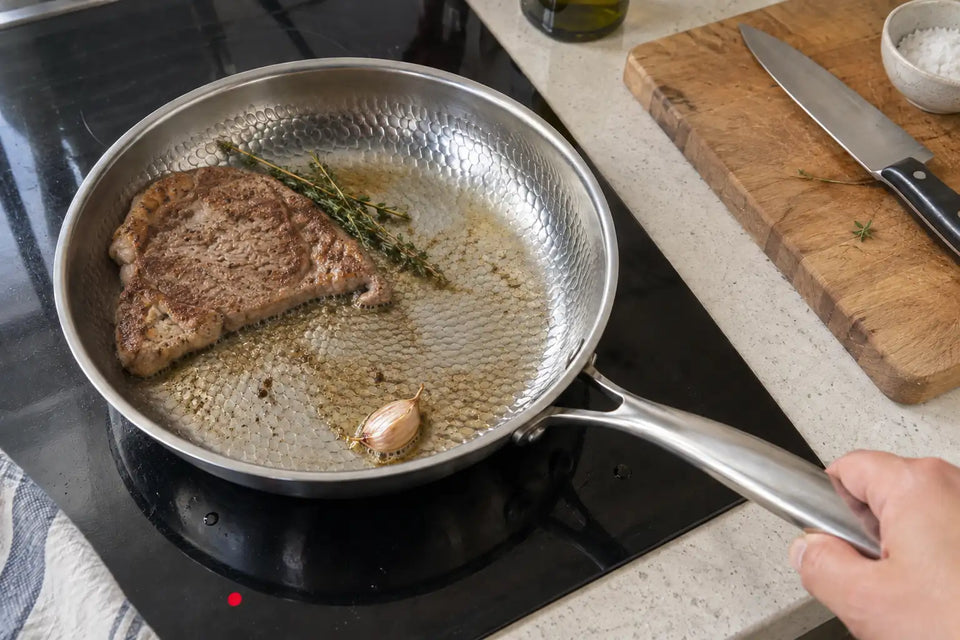 Pièce de bœuf saisie dans une poêle en titane martelé sur une plaque à induction, avec une branche de thym et une gousse d'ail, le manche tenu à la main