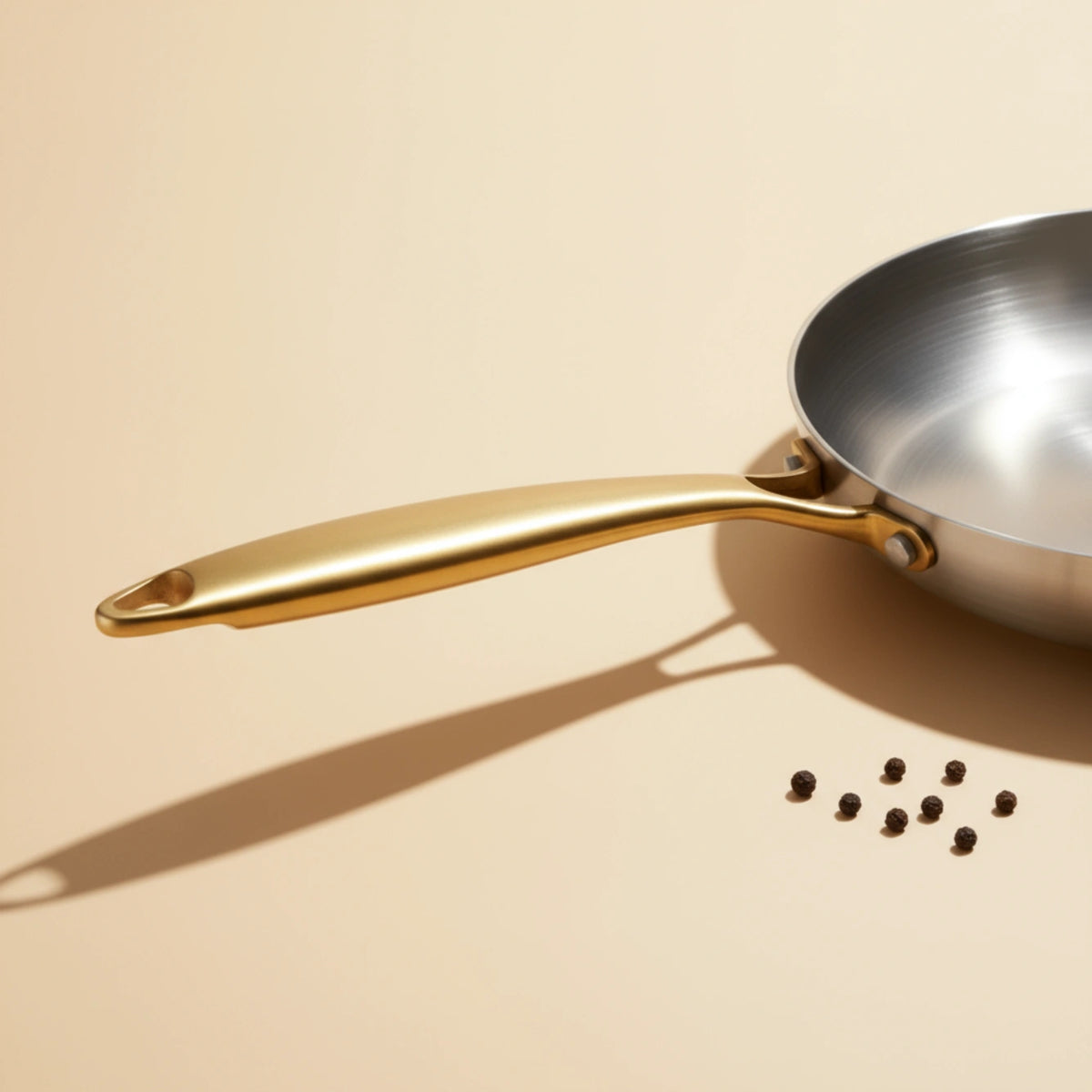 Manche doré de la poêle induction 24 cm vu de près, des grains de poivre éparpillés autour