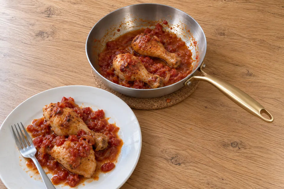 Pilons de poulet à la sauce tomate servis dans une assiette, à côté de la poêle en inox 18/10 qui a servi à les cuire