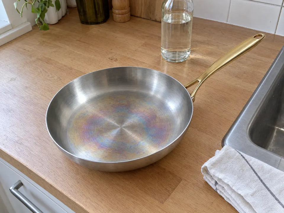 Reflets arc-en-ciel au centre de la surface de cuisson d'une poêle inox 28 cm, une bouteille de vinaigre blanc et un torchon à côté