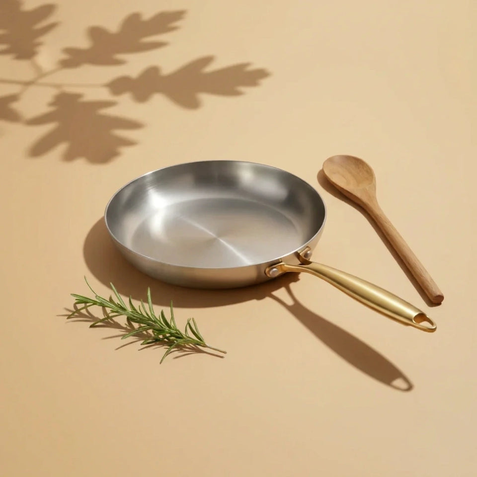 Poêle inox 28 cm vue de trois quarts sur un fond beige, une cuillère en bois et un brin de romarin à côté