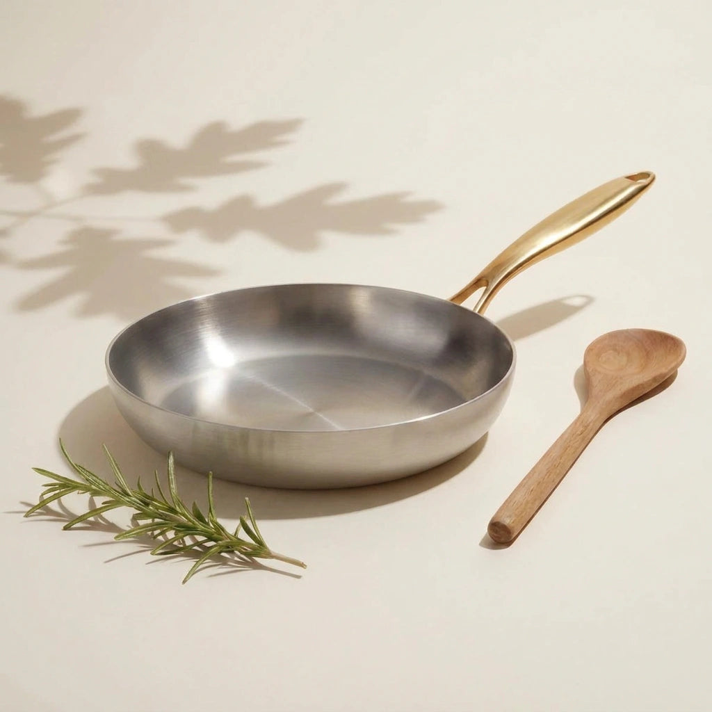 Poêle inox 30 cm vue de dessus sur un fond ivoire, sa très large surface de cuisson brossée, une cuillère en bois à côté