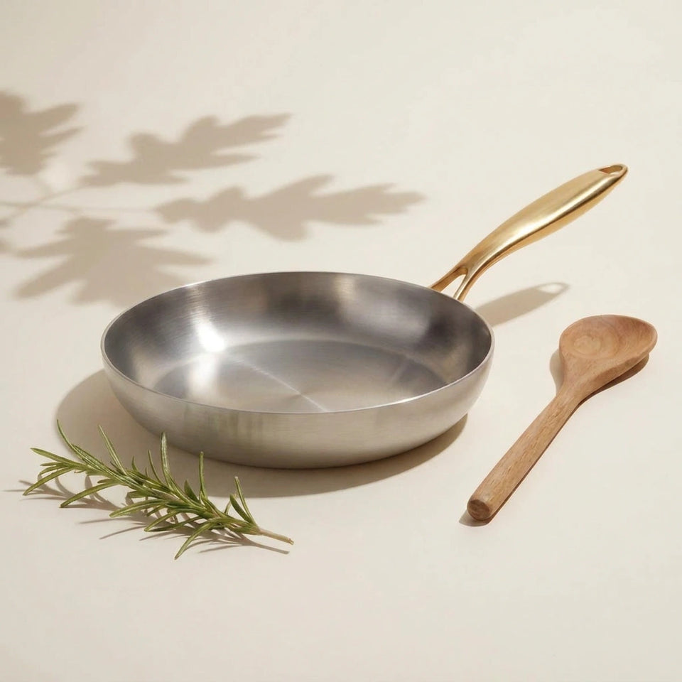 Poêle inox 30 cm vue de dessus sur un fond ivoire, sa très large surface de cuisson brossée, une cuillère en bois à côté