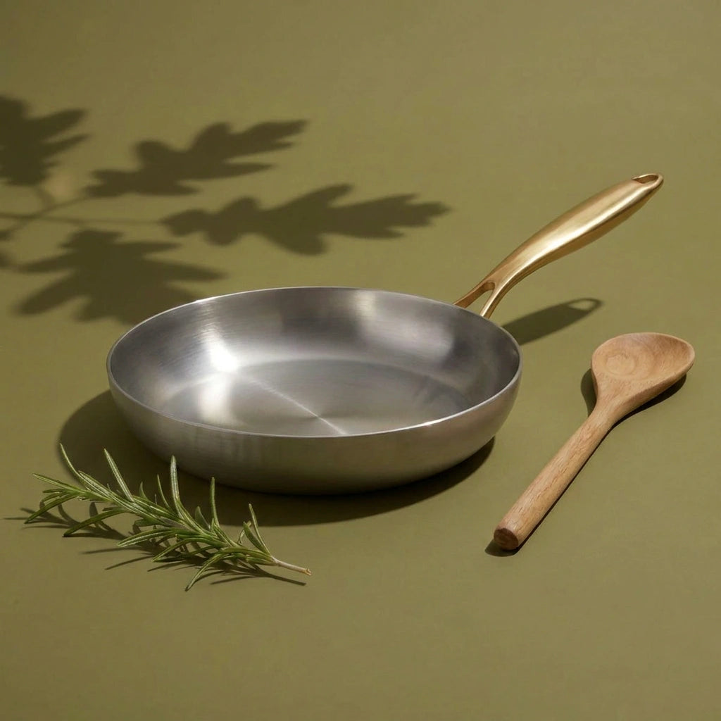 Poêle tous feux 22 cm vue de face, sa surface de cuisson brossée et son manche doré, près d'une cuillère en bois