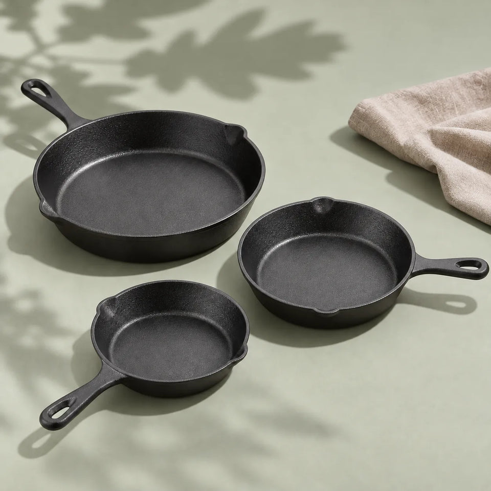 Set de 3 poêles en fonte : les trois poêles de 16, 20 et 26 cm sur un fond vert sauge, à côté d'un torchon en lin