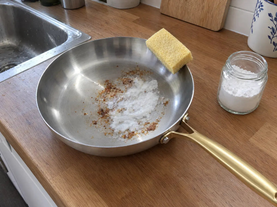 Pâte de bicarbonate de soude sur des traces de cuisson dans une poêle en inox, une éponge douce posée sur le bord