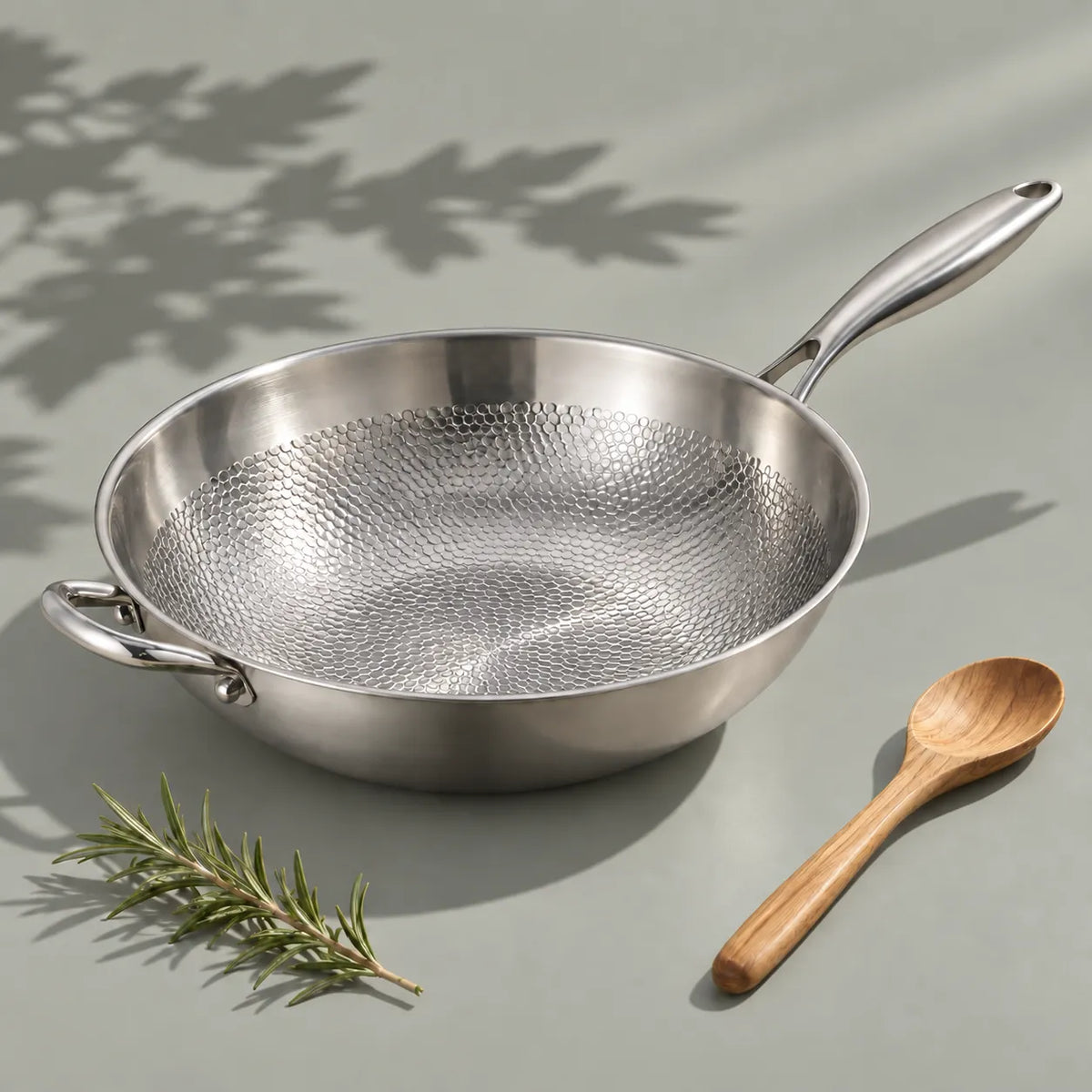 Wok en titane martelé 32 cm vu de trois quarts, sa surface de cuisson alvéolée, une cuillère en bois et du romarin à côté